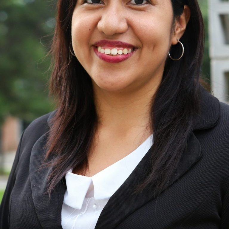 Femme souriante portant un blazer noir et une chemise blanche, arrière-plan flou.
