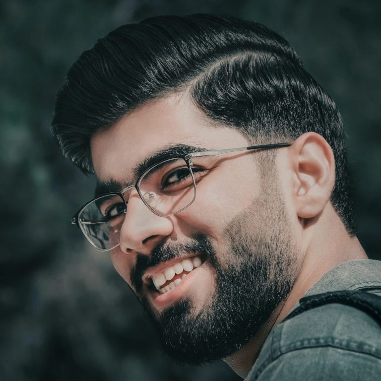 Un homme souriant avec des lunettes, cheveux foncés et une barbe, sur un fond flou.