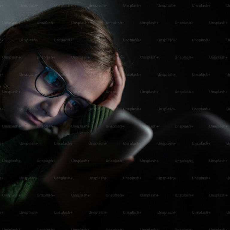 cyber harcèlement Une jeune personne avec des lunettes, absorbée par son téléphone dans une pièce sombre.