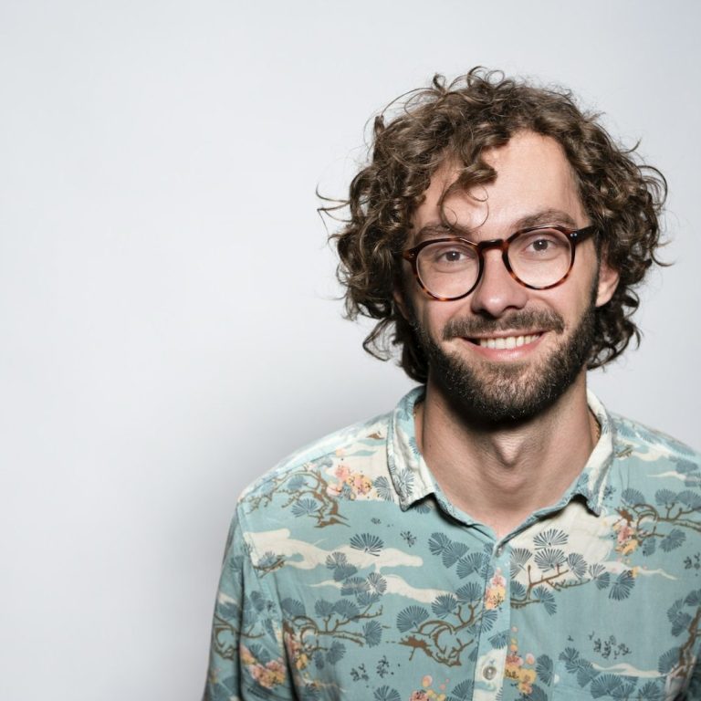Homme souriant avec des lunettes, portant une chemise à motifs colorés.